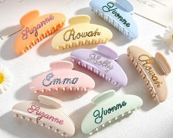 May include: A collection of eight pastel-colored plastic hair claw clips, each featuring a name spelled out in small, sparkling rhinestones. The clips come in shades of orange, yellow, light blue, lavender, pink, beige, and mint green.