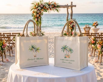 May include: Two off-white canvas tote bags with tan handles. The left bag features a palm tree illustration with the text Emily & Michael 2026. The right bag features a palm tree and tropical flower illustration with the text CABO SAN LUCAS.