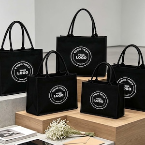 Könnte beinhalten: Fünf schwarze, rechteckige Tragetaschen in verschiedenen Größen, jede mit einer kreisförmigen weißen Grafik und dem Text "YOUR TEXT YOUR LOGO" in der Mitte. Die Taschen haben zwei stabile schwarze Griffe und ein strukturiertes Design mit flachem Boden.