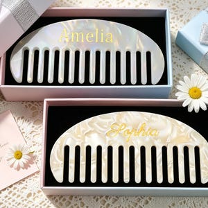 May include: Two semi-circular hair combs with a pearlescent white finish, each presented in a gift box. The top comb features the name Amelia in gold lettering, while the bottom comb displays the name Sophia in gold script. Both combs have wide teeth.