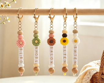 May include: Five keychains hanging in a row, each featuring a gold-toned metal clasp, a chain, and a series of beads. The beads include silicone spheres in muted tones, a flower-shaped bead, and white square letter beads spelling BLIBO, NHOBL, BALON, SLTEN, and SHTEN.