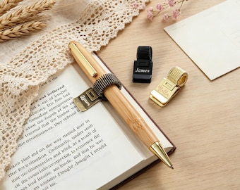 May include: A bamboo ballpoint pen with a gold-tone clip and a bronze-colored metal pen holder attached to the side. The holder is engraved with the name Liam. Two additional metal pen holders are nearby, one black engraved James and one gold engraved Stella.