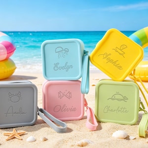 Op de afbeelding: Een stapel van vijf vierkante, pastelkleurige etuis met bijpassende polsbandjes, elk met een naam en een klein pictogram. De etuis staan op een zandstrand met schelpen, zeesterren en kleurrijke opblaasringen op de achtergrond.