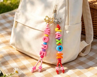 May include: Two beaded zipper pulls attached to a cream-colored backpack. One pull features pink letter beads spelling WORE with star charms, and the other features multicolored letter beads spelling JAXOM with star charms, both with silver bows.