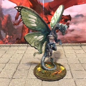 May include: A blue and green dragon miniature with butterfly wings. The dragon has a long tail and is standing on a base with green grass and mushrooms.