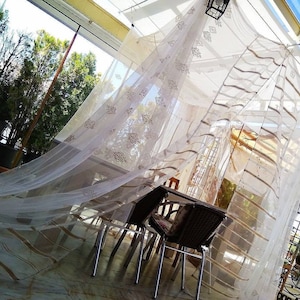 Puede incluir: Una cortina blanca y transparente con rayas marrones cuelga sobre un juego de comedor de patio. La cortina se mueve con el viento, creando un ambiente caprichoso y aireado.