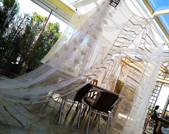 May include: A white sheer curtain with brown stripes hangs over a patio dining set. The curtain is blowing in the wind, creating a whimsical and airy atmosphere.