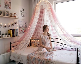 May include: A bedroom with a bed featuring a pink and white canopy with fairy lights. A woman in a floral dress sits on the bed, holding a book. The room includes a bookshelf with books, photos, and decorative items.