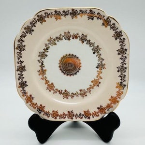 May include: A square, vintage-style plate with a cream-colored surface and a gold rim. The plate features a floral design in brown and gold around the edge and center. The plate is displayed on a black stand.