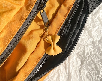 May include: Mustard yellow fanny pack with black zippers and a pasta-shaped charm. The charm is light yellow and hangs from the zipper. The background is a white, textured surface. The bag's design is visible.
