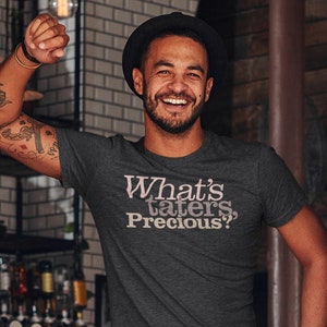 May include: A man wearing a dark grey t-shirt with the text "What's taters Precious?" in pink lettering. He is standing in a doorway with a wooden door frame. The text "SOMETHING flourished" is written in white on a black background with white rays emanating from the text.