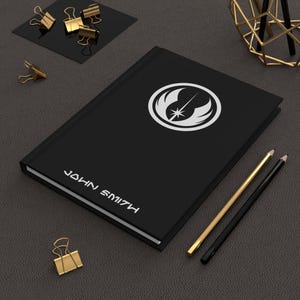 Puede incluir: Diario negro con un logotipo blanco y el nombre "JOHN SMITH" impreso en blanco. El diario está sobre una superficie gris oscuro con lápices dorados y negros, y clips dorados.