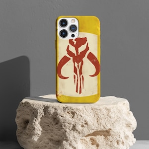 May include: A yellow iPhone case with a red and white Mandalorian symbol. The case has a distressed look and is on a light brown rock.