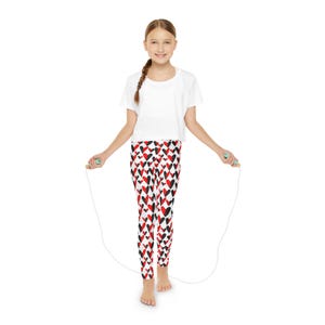 May include: A young person wearing a white t-shirt and red, black, and white heart patterned leggings is jumping rope. The rope is white with green handles.