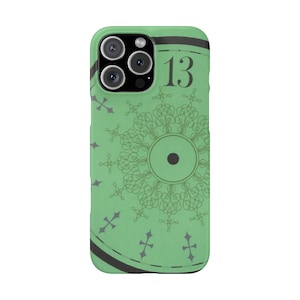 May include: A green phone case featuring a decorative design with the number 13. The case has a black border and a circular pattern with a central dot. Small cross-shaped accents are scattered around the design.