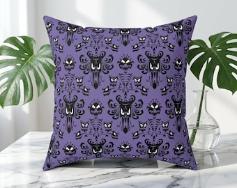 May include: Square purple decorative pillow with a repeating black pattern of stylized haunted house faces. The pillow is on a white marble surface, with green plants in the background.