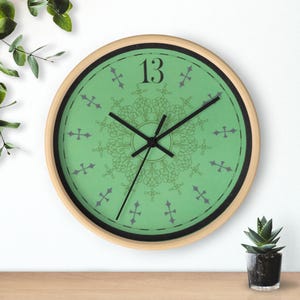 May include: A round wall clock with a light wood frame. The clock face is green with black hands and the number 13. It features a decorative, symmetrical design in the center and gray cross-shaped markers around the edge.