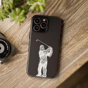 May include: Black phone case featuring a white illustration of Chewbacca playing golf. The design shows Chewbacca in a golf swing pose with a golf club. The phone case is on a wooden surface.