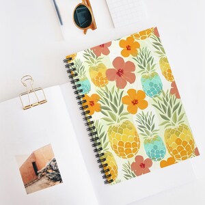 May include: A spiral-bound notebook with a colourful pattern of yellow and turquoise pineapples, orange flowers, and green leaves on a cream background. The notebook is open, revealing blank pages, with a gold paper clip and sunglasses.