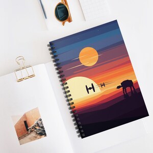 May include: Spiral-bound notebook featuring a sunset scene with two suns, a silhouette of a walker, and two TIE fighters. The cover design has a gradient of orange, yellow, and purple hues. The notebook is open on a white surface.