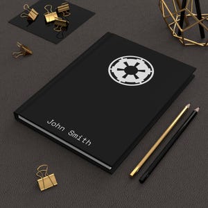 Könnte beinhalten: Schwarzes Notizbuch mit einem weißen Imperial-Symbol und dem Namen "John Smith" auf dem Einband. Goldene und schwarze Bleistifte sowie goldene Büroklammern sind um das Notizbuch auf einer dunkelgrauen Oberfläche angeordnet.