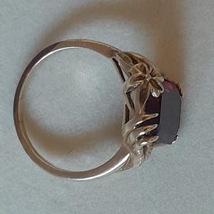May include: A gold-toned ring featuring a deep red, rectangular-cut gemstone set within a decorative metal band. The setting includes sculpted floral and leaf-like metal details that extend from the band to frame the central dark red stone.