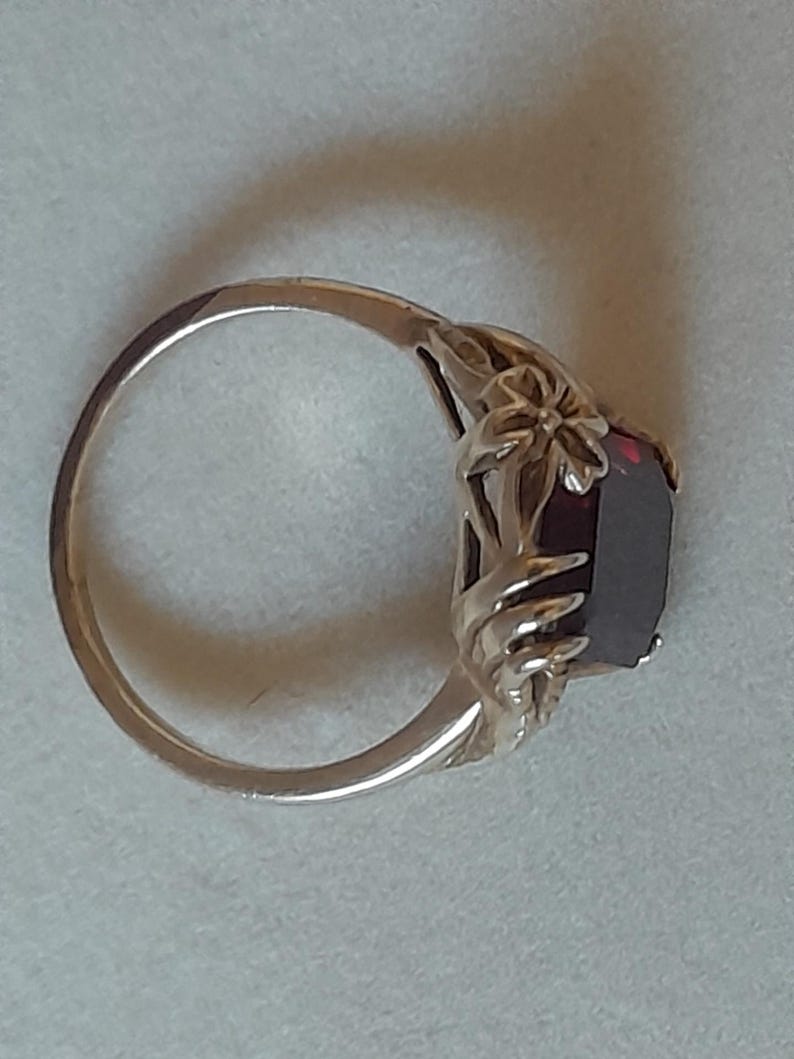 May include: A gold-toned ring featuring a deep red, rectangular-cut gemstone set within a decorative metal band. The setting includes sculpted floral and leaf-like metal details that extend from the band to frame the central dark red stone.