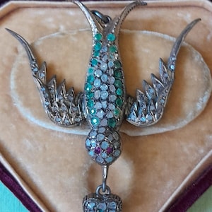 Op de afbeelding: Een vintage zilverkleurige vogelhanger met smaragdgroene en heldere edelsteenaccenten. De vogel heeft uitgespreide vleugels en een hartvormige hanger. De sieraden worden gepresenteerd in een met fluweel gevoerde doos.