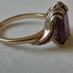 May include: A gold ring featuring a rectangular, deep red gemstone set in a decorative metal mounting. The band is smooth and polished, transitioning into textured, leaf-like metal details that frame the central stone on both sides.