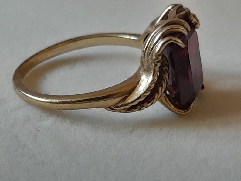 May include: A gold ring featuring a rectangular, deep red gemstone set in a decorative metal mounting. The band is smooth and polished, transitioning into textured, leaf-like metal details that frame the central stone on both sides.