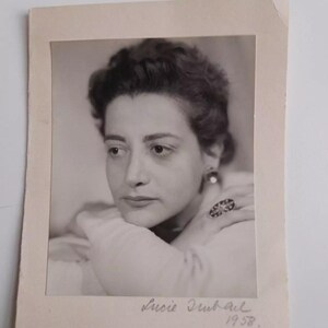 May include: Black and white photograph of a woman with short, dark hair. She is wearing a white blouse and a large ring on her finger. The photo is signed "Lucie Dubail 1958."