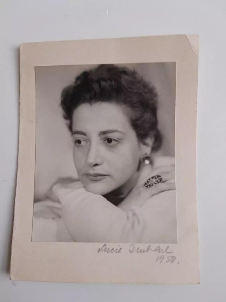 May include: Black and white photograph of a woman with short, dark hair. She is wearing a white blouse and a large ring on her finger. The photo is signed "Lucie Dubail 1958."