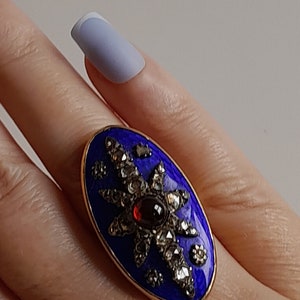 May include: A gold ring with a blue enamel oval setting. The oval is decorated with a starburst design of small diamonds and a red garnet in the center.