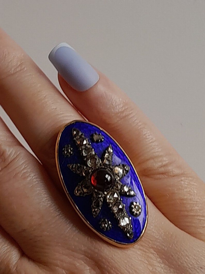 May include: A gold ring with a blue enamel oval setting. The oval is decorated with a starburst design of small diamonds and a red garnet in the center.