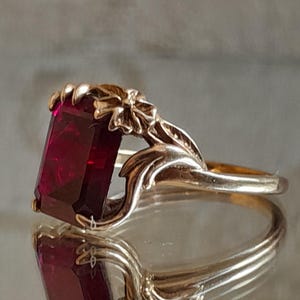 May include: A gold ring featuring a large, rectangular, deep red gemstone set in a prong setting. The band is designed with sculpted metal floral and leaf motifs that extend from the sides to frame the central stone, creating a detailed look.