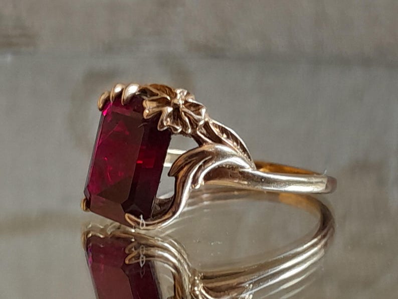 May include: A gold ring featuring a large, rectangular, deep red gemstone set in a prong setting. The band is designed with sculpted metal floral and leaf motifs that extend from the sides to frame the central stone, creating a detailed look.