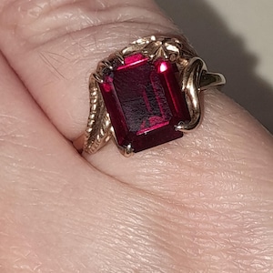 May include: A gold ring featuring a large, rectangular, deep red gemstone set in a prong setting. The band of the ring displays a textured, rope-like design on the sides, leading up to the central stone which is held by four metal prongs.