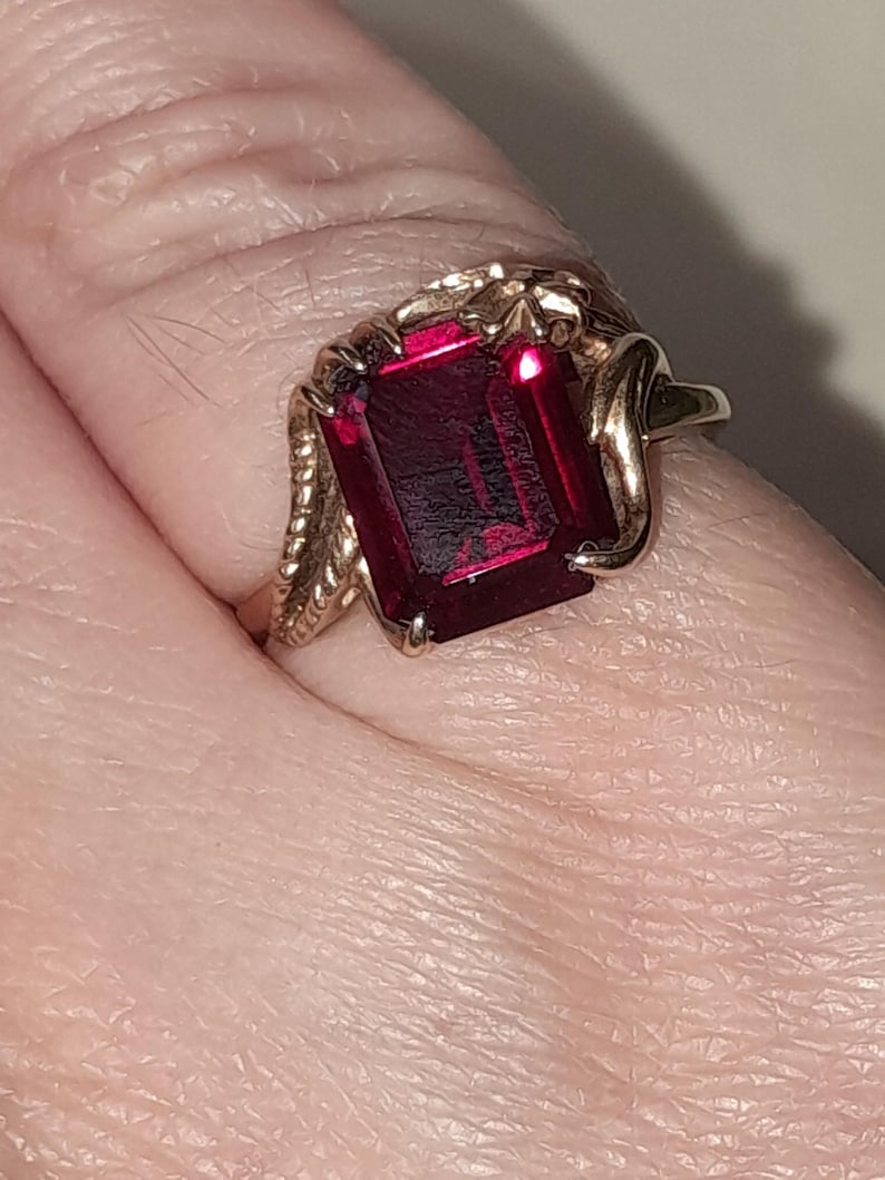 May include: A gold ring featuring a large, rectangular, deep red gemstone set in a prong setting. The band of the ring displays a textured, rope-like design on the sides, leading up to the central stone which is held by four metal prongs.