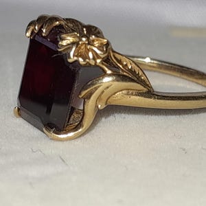 May include: A gold ring featuring a rectangular, deep red gemstone set in a band decorated with sculpted metal floral and leaf motifs. The setting includes prongs that secure the stone, while the shank displays detailed botanical engravings.