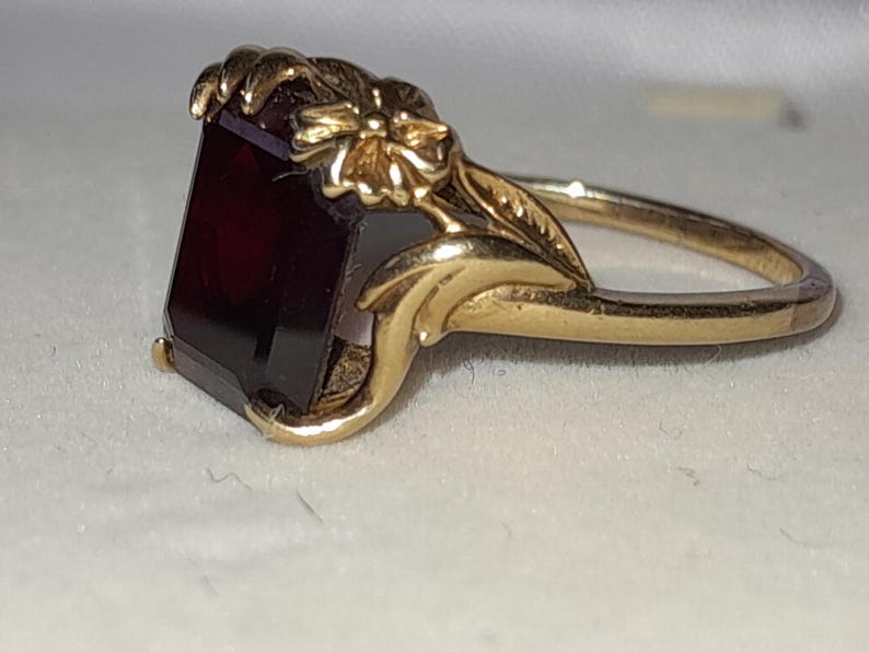 May include: A gold ring featuring a rectangular, deep red gemstone set in a band decorated with sculpted metal floral and leaf motifs. The setting includes prongs that secure the stone, while the shank displays detailed botanical engravings.