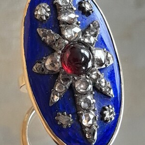 May include: An oval-shaped ring with a blue enamel setting. The ring features a starburst design with a red gemstone in the center and smaller diamonds surrounding it.
