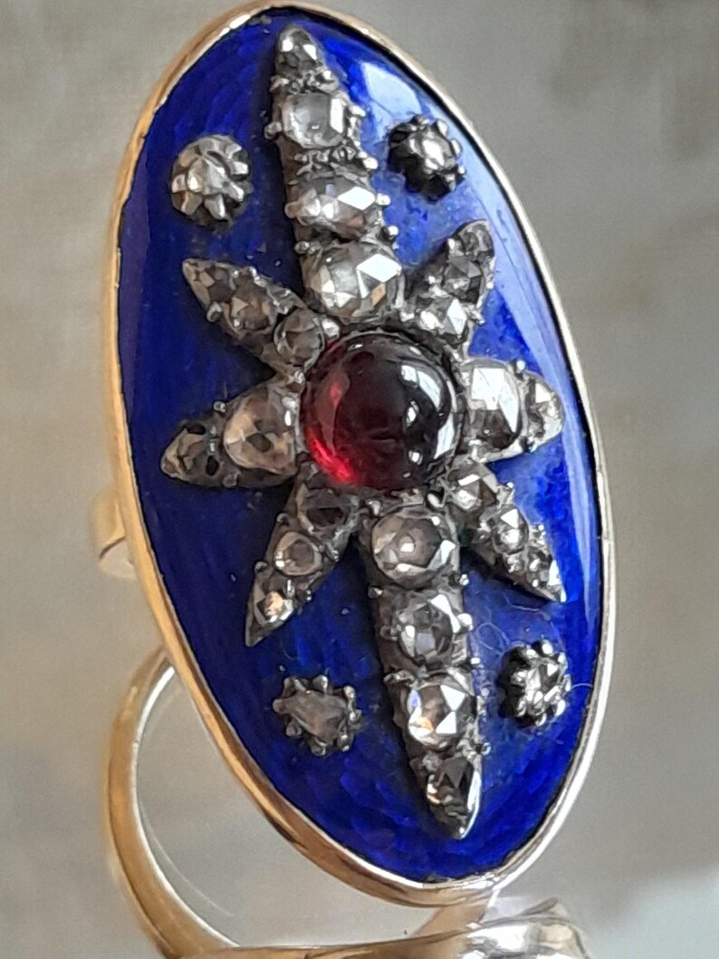 May include: An oval-shaped ring with a blue enamel setting. The ring features a starburst design with a red gemstone in the center and smaller diamonds surrounding it.