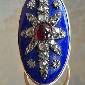 May include: A vintage ring with a blue enamel setting featuring a starburst design with a red gemstone in the center and small diamonds around it.