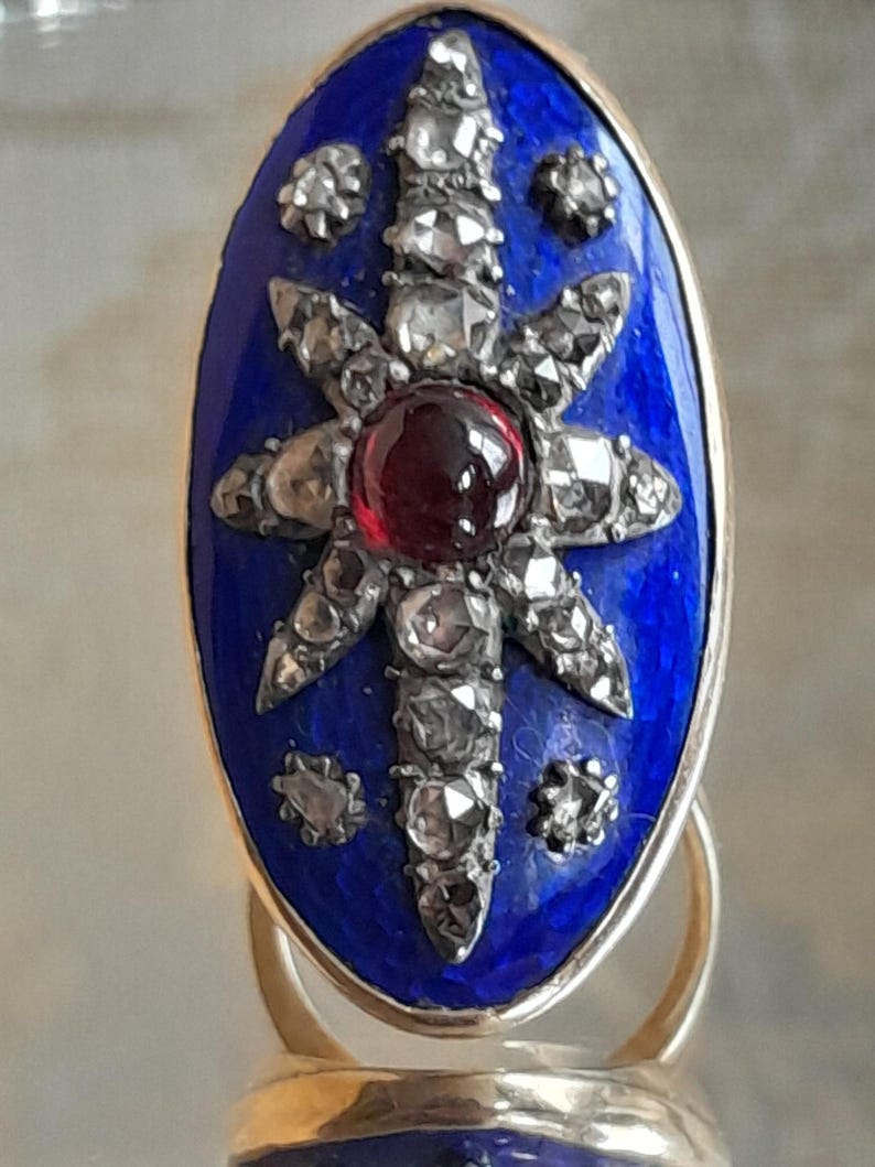 May include: A vintage ring with a blue enamel setting featuring a starburst design with a red gemstone in the center and small diamonds around it.