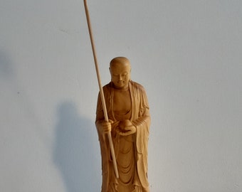 May include: A wooden statue of a Buddhist monk holding a small orb in his left hand and a staff with a decorative top in his right hand. The monk is standing on a lotus base.
