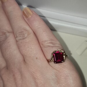 May include: A gold ring featuring a rectangular, emerald-cut deep red gemstone set in the center. The metal band displays a textured, vine-like or leaf-inspired design on both sides of the central stone, creating a detailed and ornate appearance.
