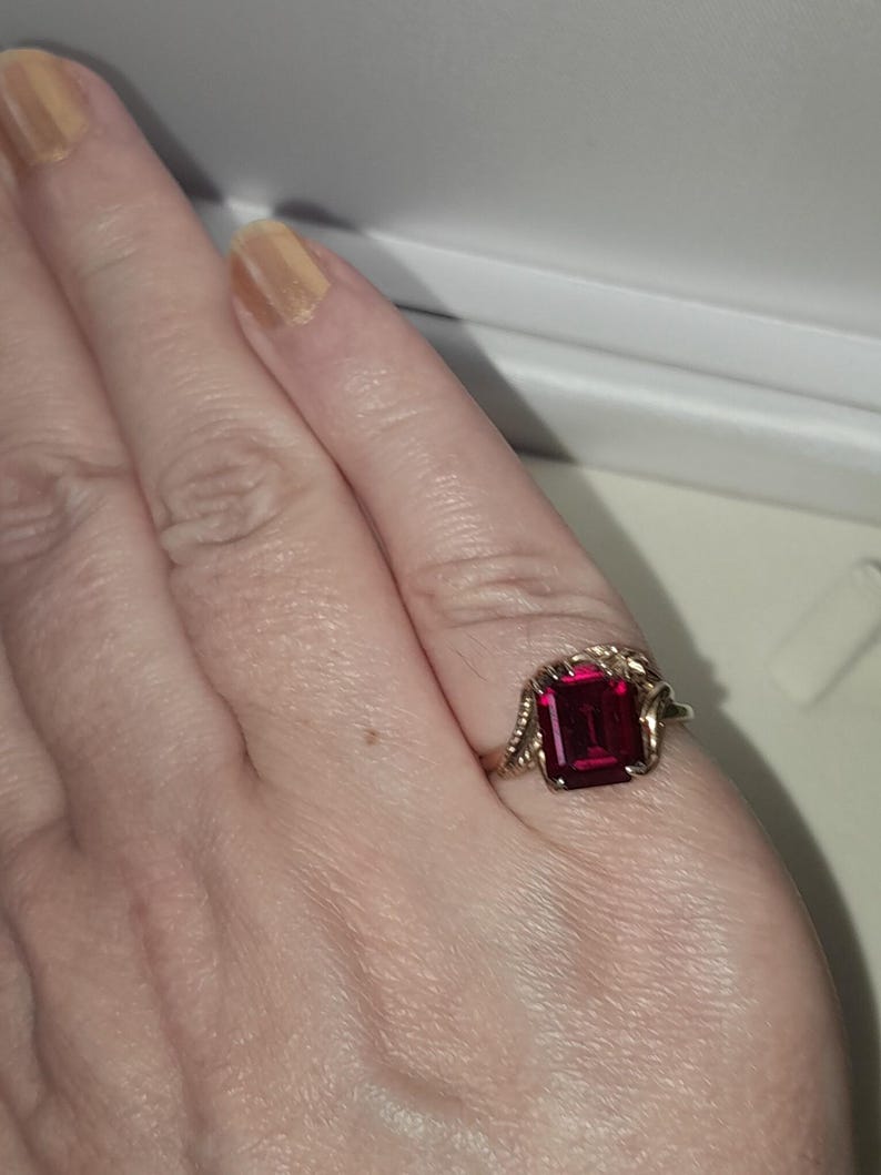 May include: A gold ring featuring a rectangular, emerald-cut deep red gemstone set in the center. The metal band displays a textured, vine-like or leaf-inspired design on both sides of the central stone, creating a detailed and ornate appearance.