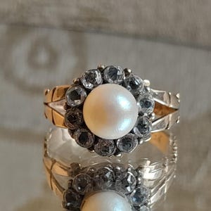 May include: A gold ring featuring a central round white pearl surrounded by a halo of ten small, clear, faceted stones. The band has a split design on the sides, and the entire piece is reflected on a smooth, shiny surface below the ring.
