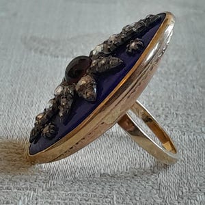 May include: A gold ring with a blue enamel oval top set with a dark red gemstone and small diamonds. The ring is on a white fabric background.