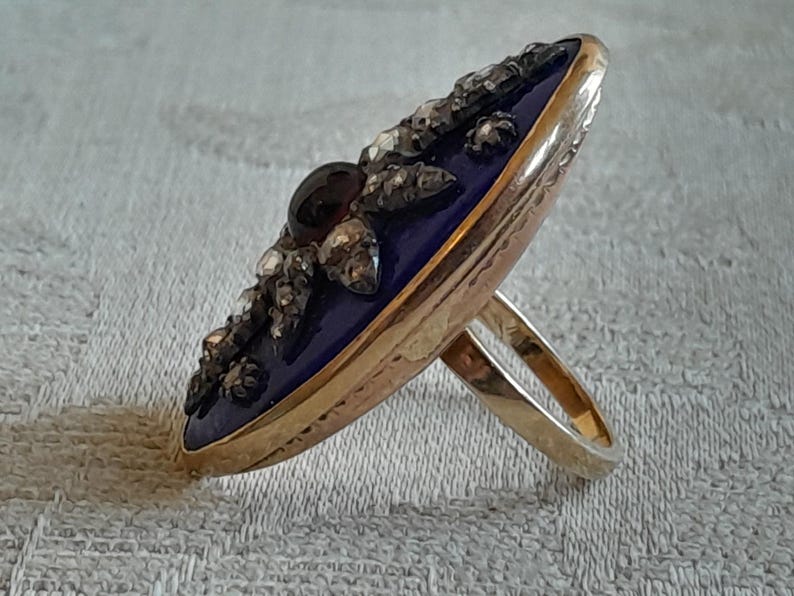 May include: A gold ring with a blue enamel oval top set with a dark red gemstone and small diamonds. The ring is on a white fabric background.